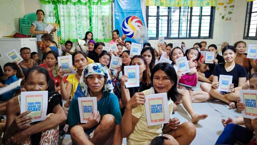 A diverse group of people, mostly women, sit closely together in a bright room, holding booklets. They appear attentive and engaged, conveying a sense of community.