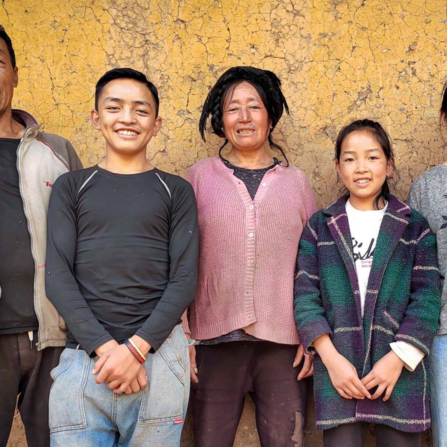 A family of five posing in front of a textured, earthy yellow wall. They are smiling warmly, dressed in casual clothes, conveying a joyful and relaxed mood.