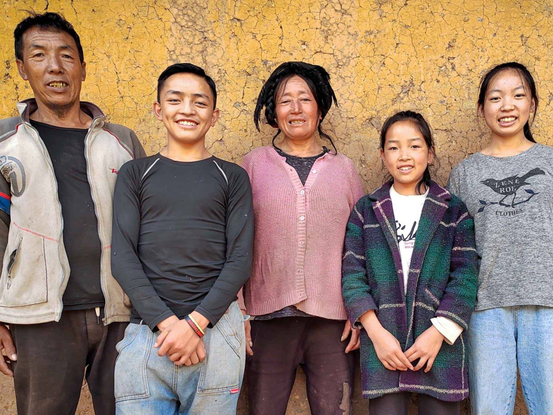 A family of five posing in front of a textured, earthy yellow wall. They are smiling warmly, dressed in casual clothes, conveying a joyful and relaxed mood.