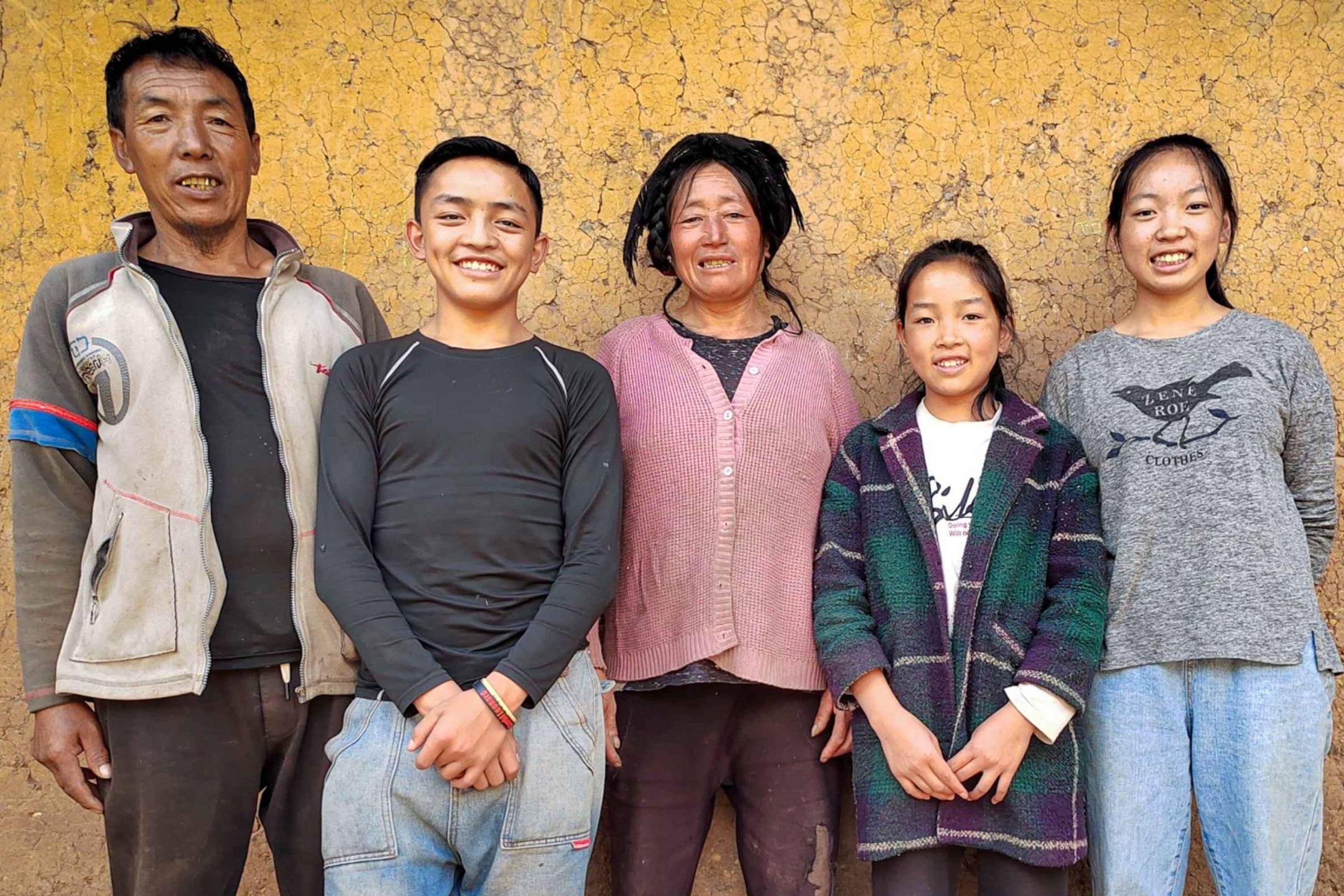 A family of five posing in front of a textured, earthy yellow wall. They are smiling warmly, dressed in casual clothes, conveying a joyful and relaxed mood.