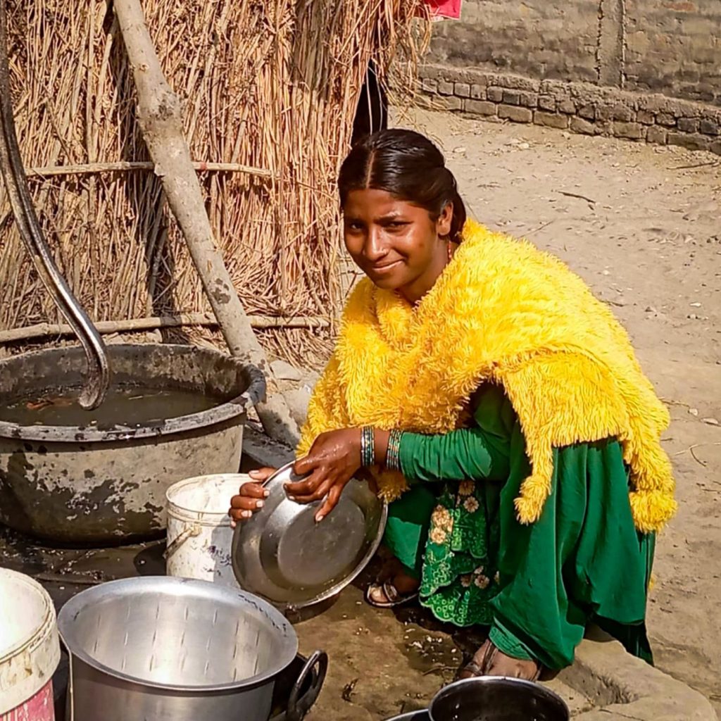 A woman in a bright yellow shawl and green dress smiles while washing dishes outdoors. She holds a pot beside a water tub, with a rustic backdrop.