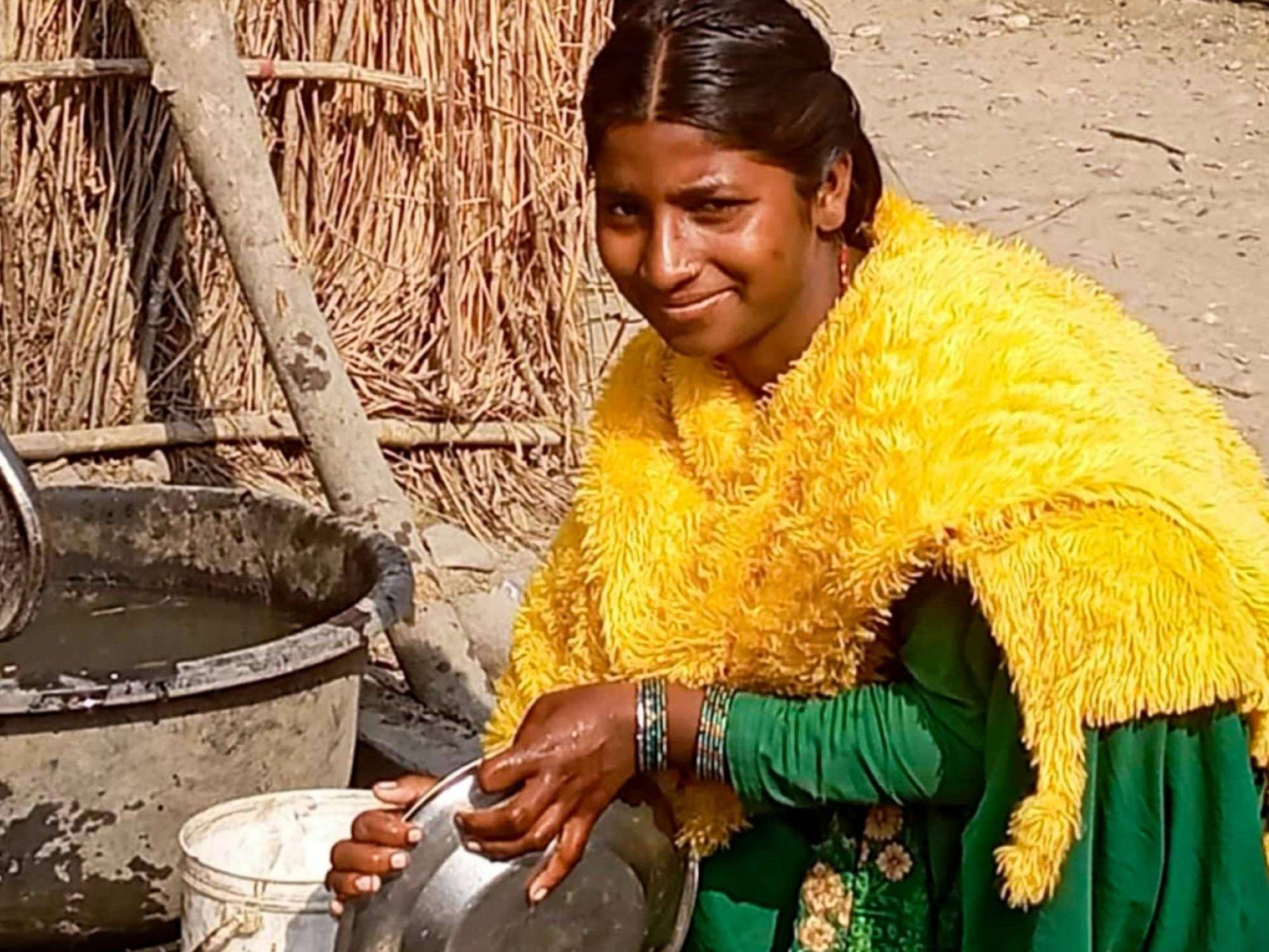 A woman in a bright yellow shawl and green dress smiles while washing dishes outdoors. She holds a pot beside a water tub, with a rustic backdrop.