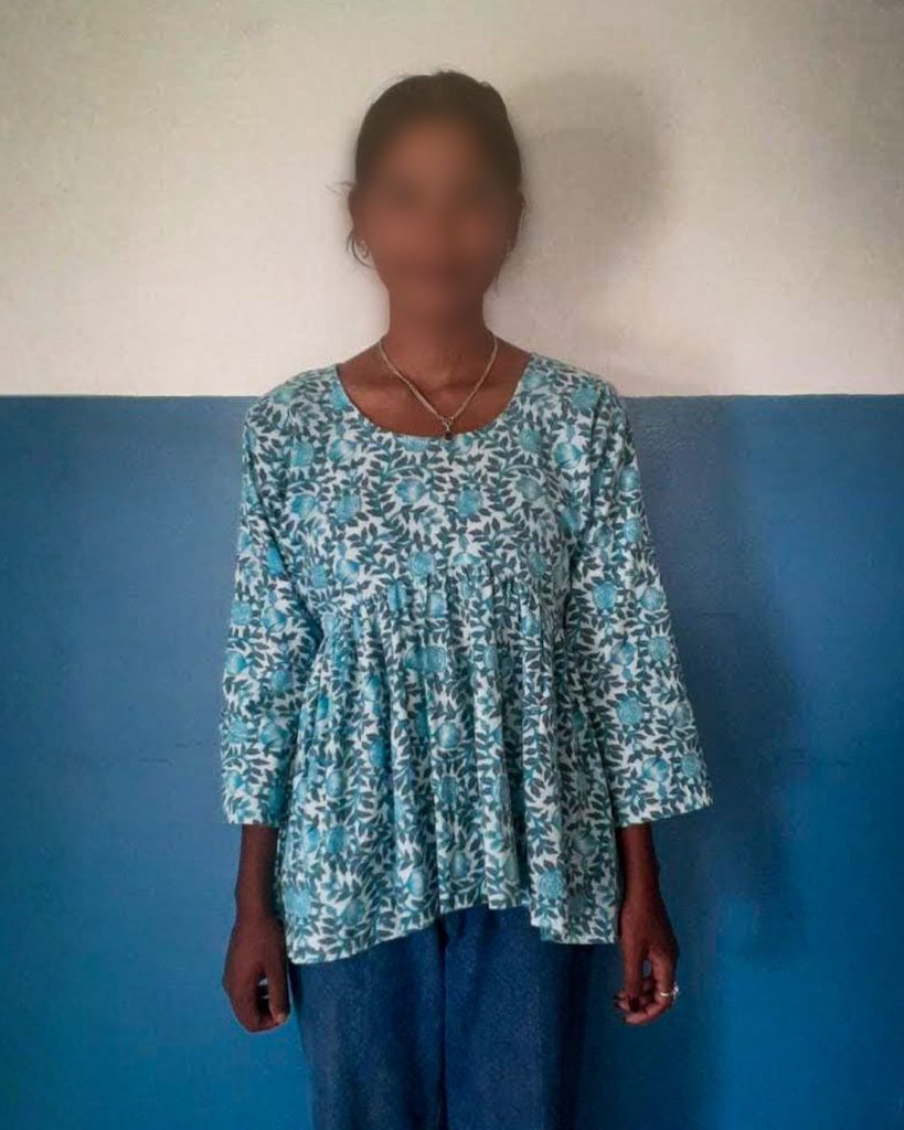 A woman stands against a plain wall, wearing a blue patterned top and jeans. Her face is blurred for privacy.