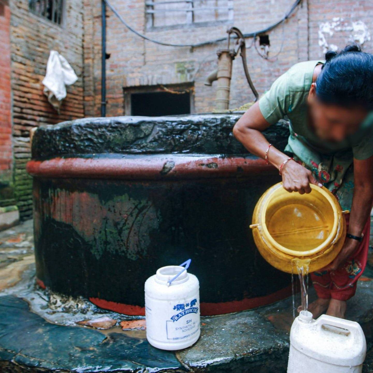 A woman in a green shirt pours water from a well into a container in a brick courtyard. The scene conveys a sense of daily life and tradition.
