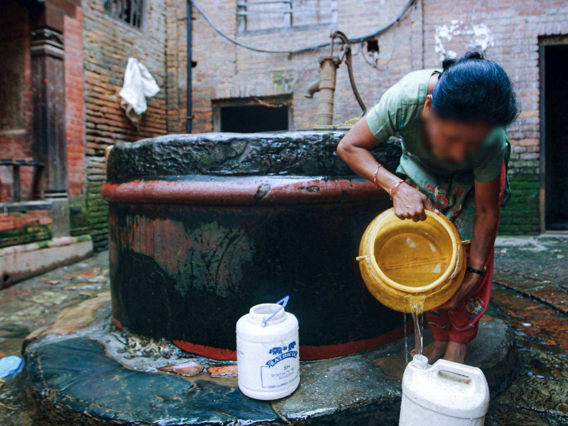 A woman in a green shirt pours water from a well into a container in a brick courtyard. The scene conveys a sense of daily life and tradition.