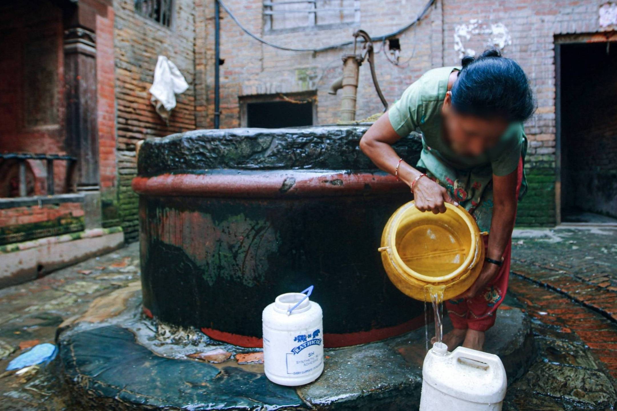 A woman in a green shirt pours water from a well into a container in a brick courtyard. The scene conveys a sense of daily life and tradition.