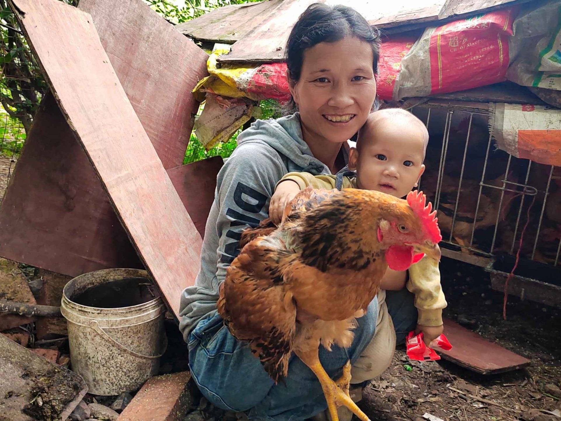 A woman and a baby smile while holding a large brown chicken in front of a rustic coop. The scene is outdoors, exuding warmth and rural charm.