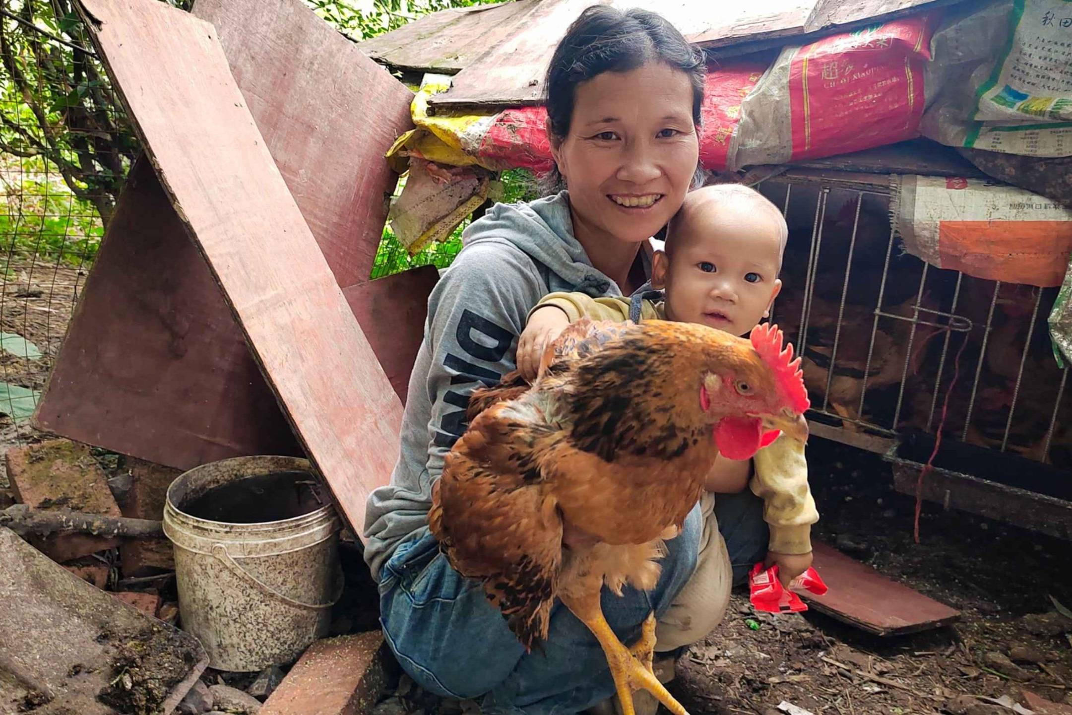 A woman and a baby smile while holding a large brown chicken in front of a rustic coop. The scene is outdoors, exuding warmth and rural charm.