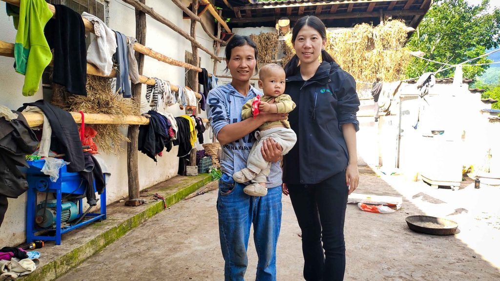A smiling person holds a baby, standing beside another person outdoors. Clothes dry on racks against a rustic building with hay and greenery.