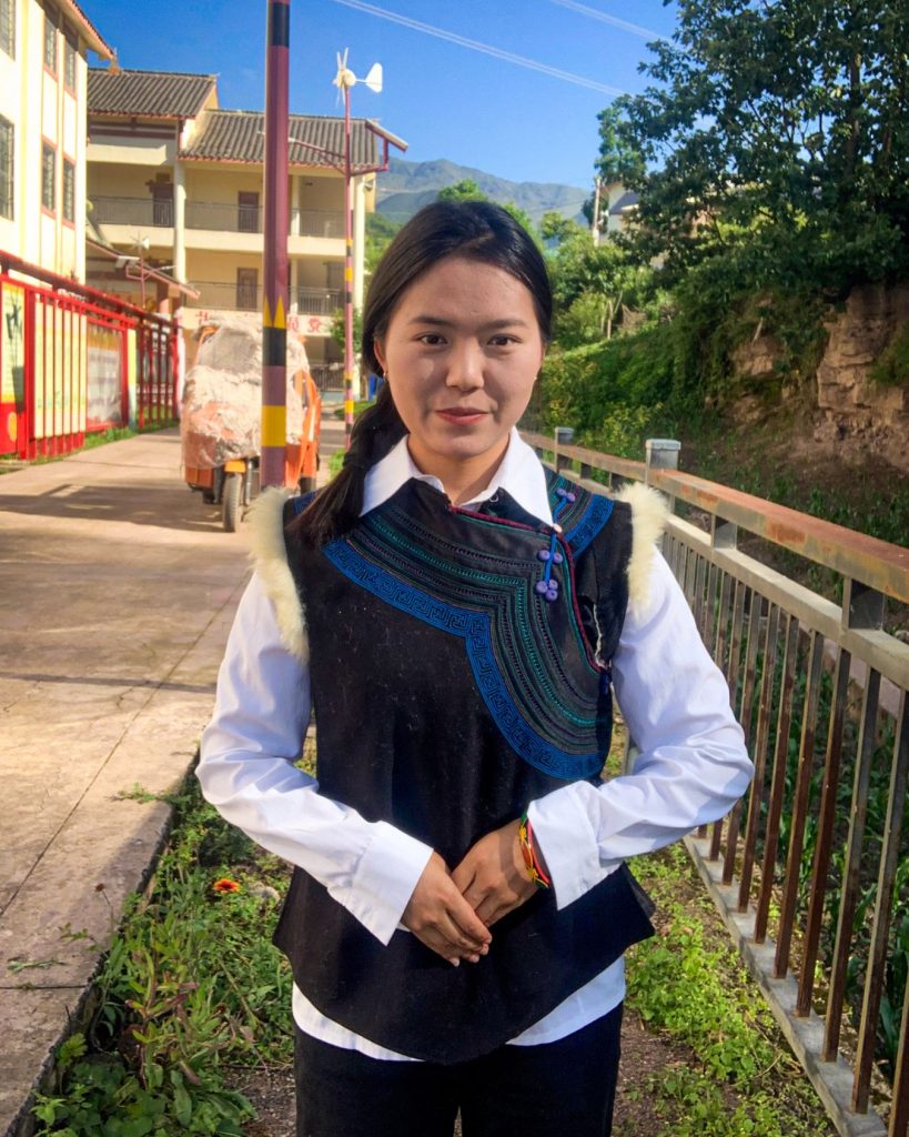 A young woman smiles warmly in colorful traditional attire, with intricate embroidery, standing in a sunlit village street flanked by greenery and buildings.