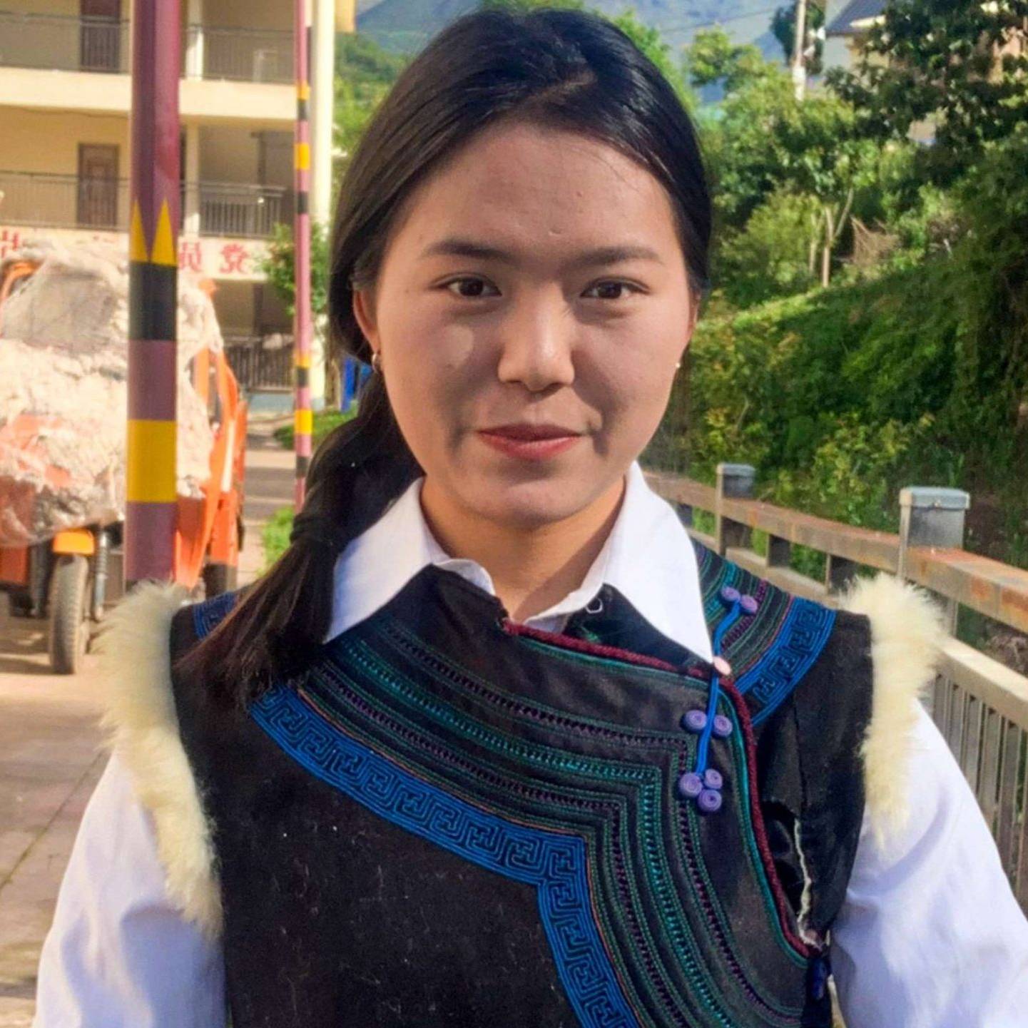 A young woman smiles warmly in colorful traditional attire, with intricate embroidery, standing in a sunlit village street flanked by greenery and buildings.