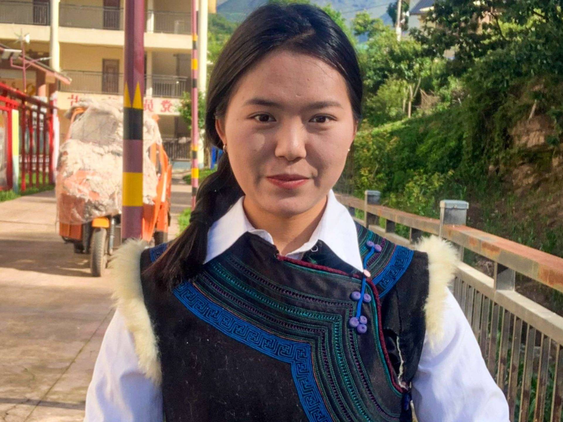 A young woman smiles warmly in colorful traditional attire, with intricate embroidery, standing in a sunlit village street flanked by greenery and buildings.