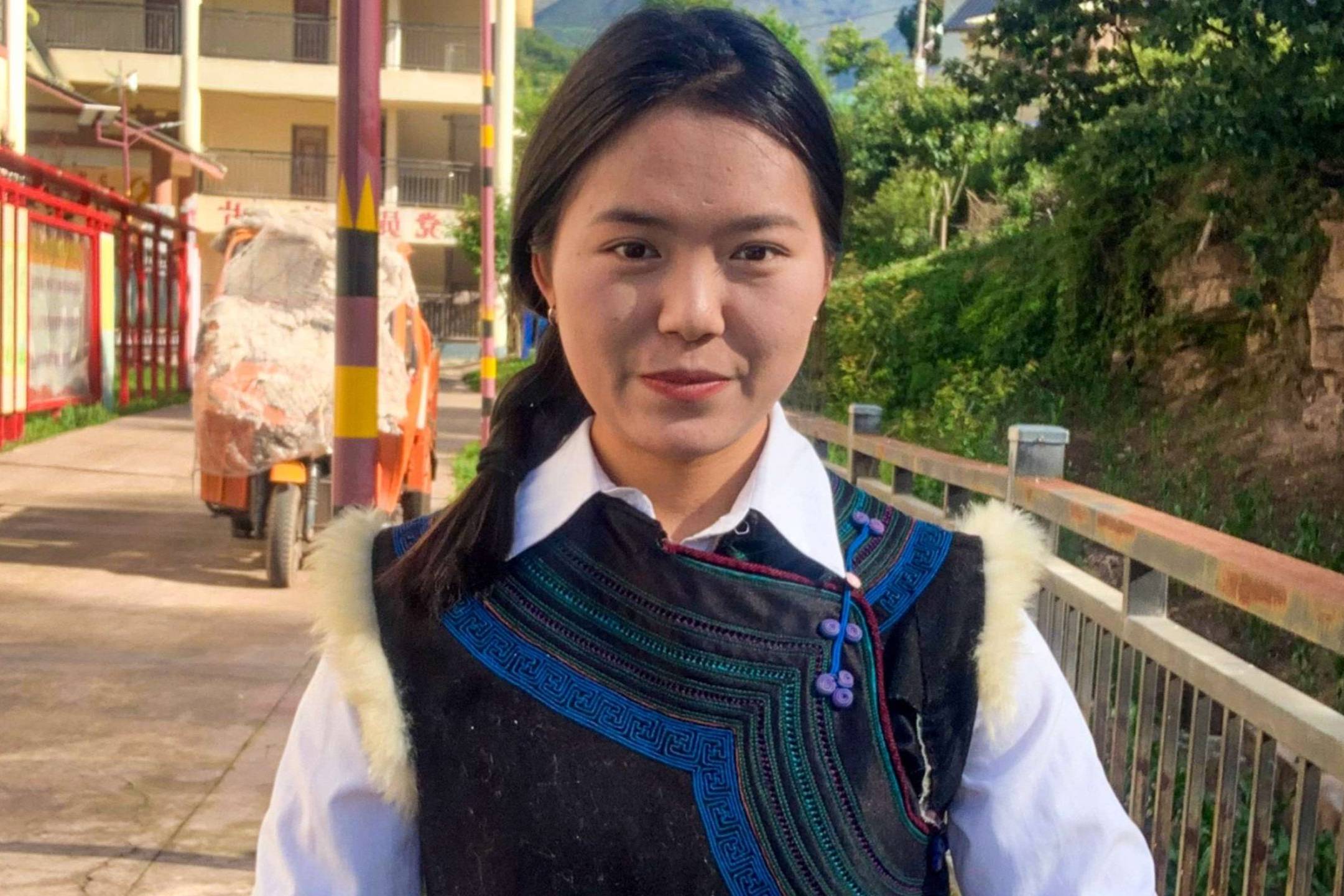 A young woman smiles warmly in colorful traditional attire, with intricate embroidery, standing in a sunlit village street flanked by greenery and buildings.
