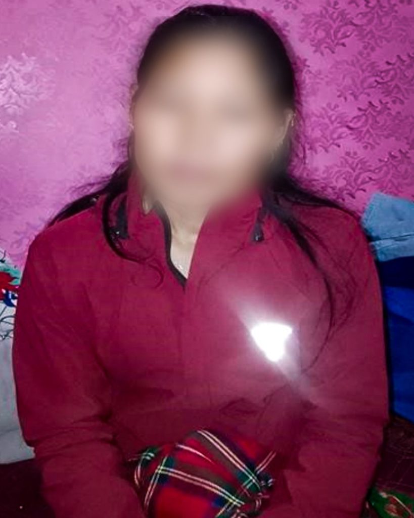 A person in a red jacket sits against a pink patterned wall, holding a plaid scarf. Her face blurred for privacy.