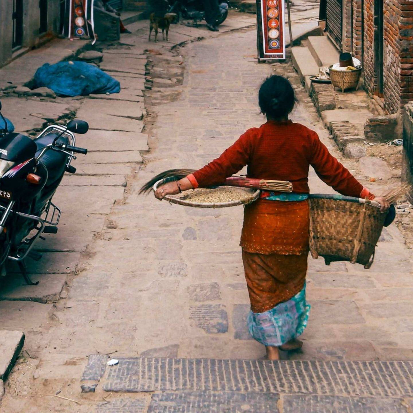 A woman in traditional attire walks down a narrow, cobbled street carrying a wicker basket and a tray. A motorbike is parked nearby.