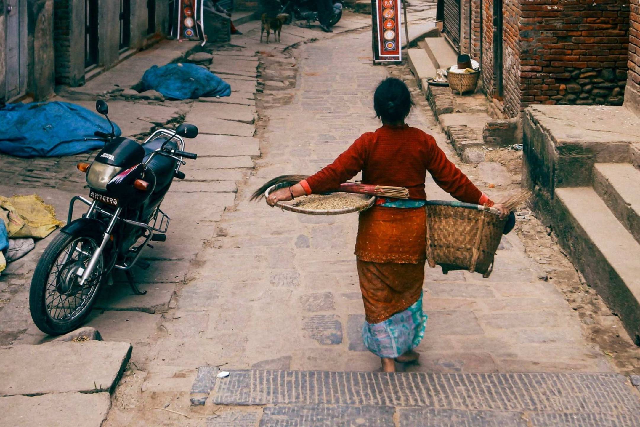 A woman in traditional attire walks down a narrow, cobbled street carrying a wicker basket and a tray. A motorbike is parked nearby.