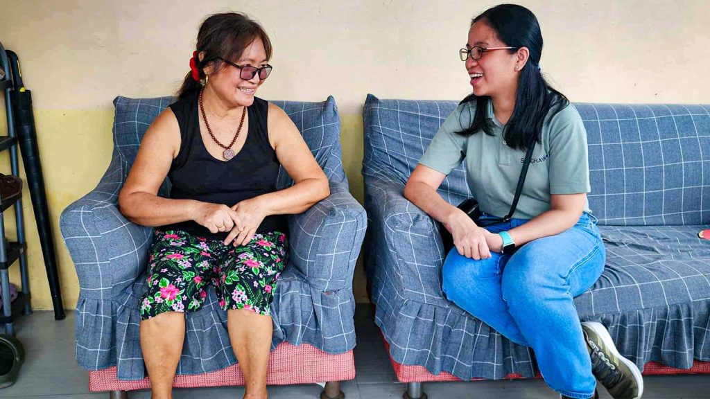 Two women sitting on a plaid-covered sofa, engaged in a cheerful conversation. One wears floral pants and glasses; the other in jeans and a green shirt.