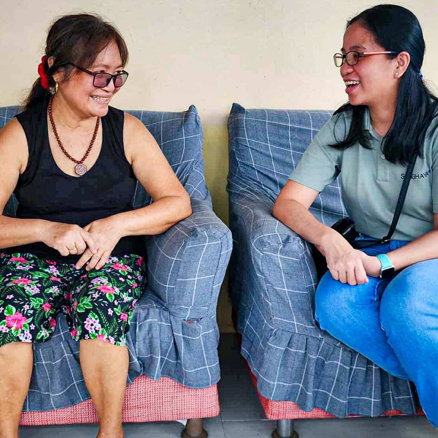 Two women sitting on a plaid-covered sofa, engaged in a cheerful conversation. One wears floral pants and glasses; the other in jeans and a green shirt.