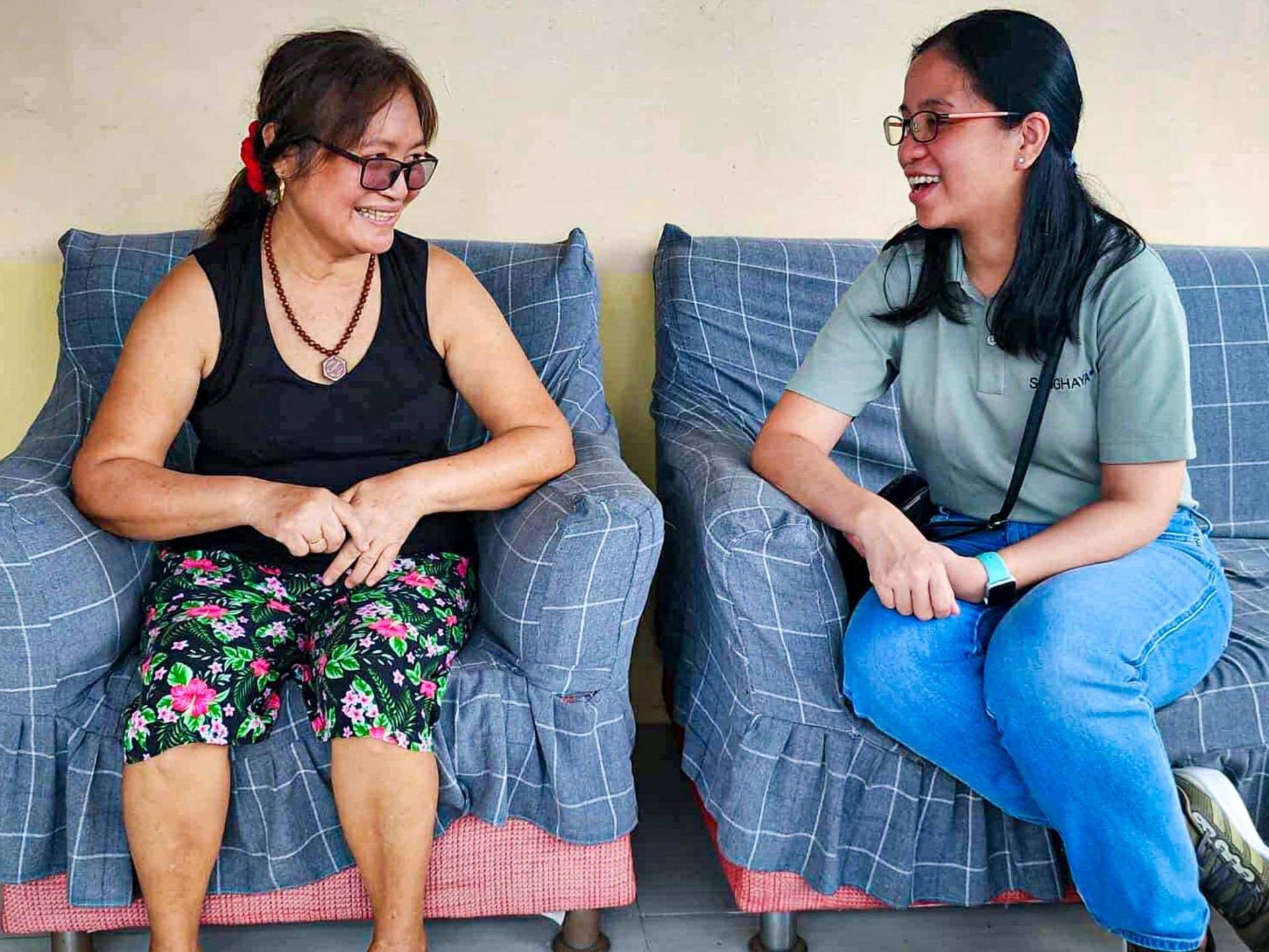 Two women sitting on a plaid-covered sofa, engaged in a cheerful conversation. One wears floral pants and glasses; the other in jeans and a green shirt.