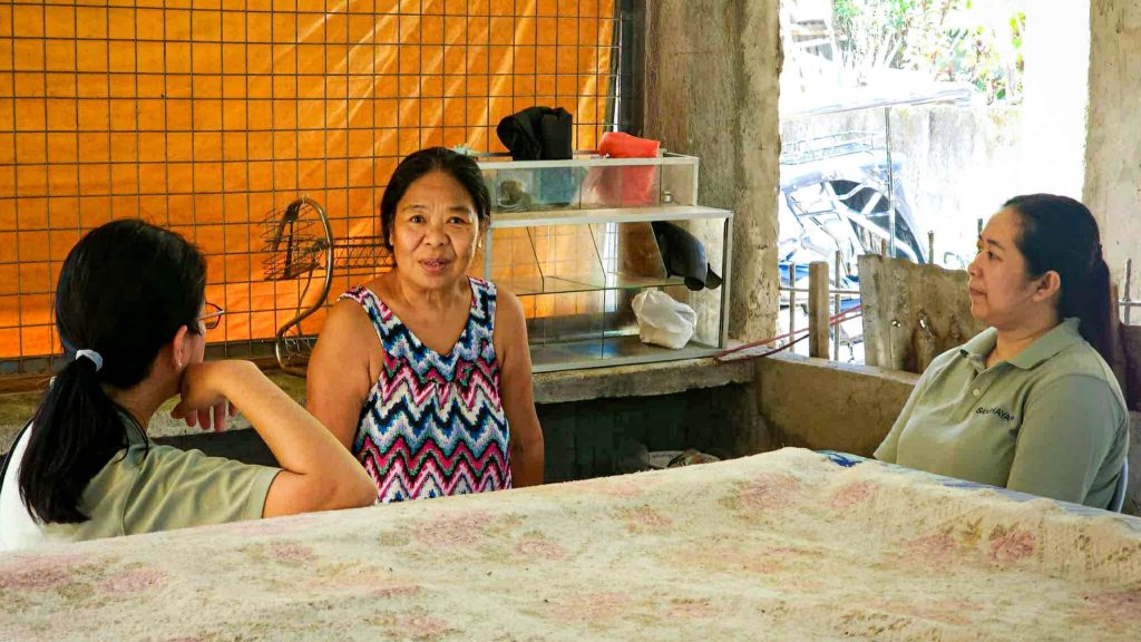 Three women interact in a rustic setting with a table covered in a floral cloth.