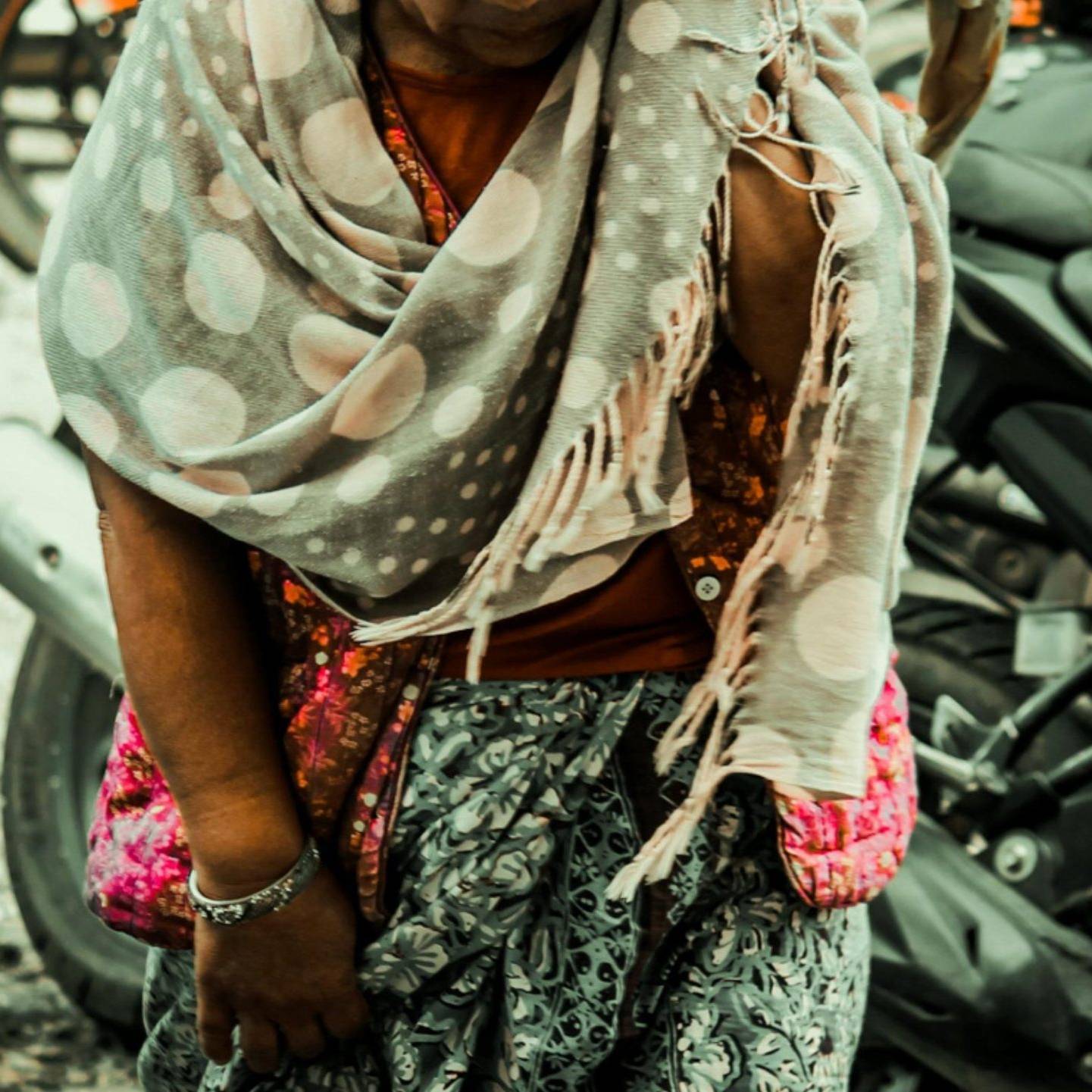 An elderly person, wrapped in a polka dot scarf and patterned clothing, leans forward while walking near parked motorcycles, conveying a sense of struggle.