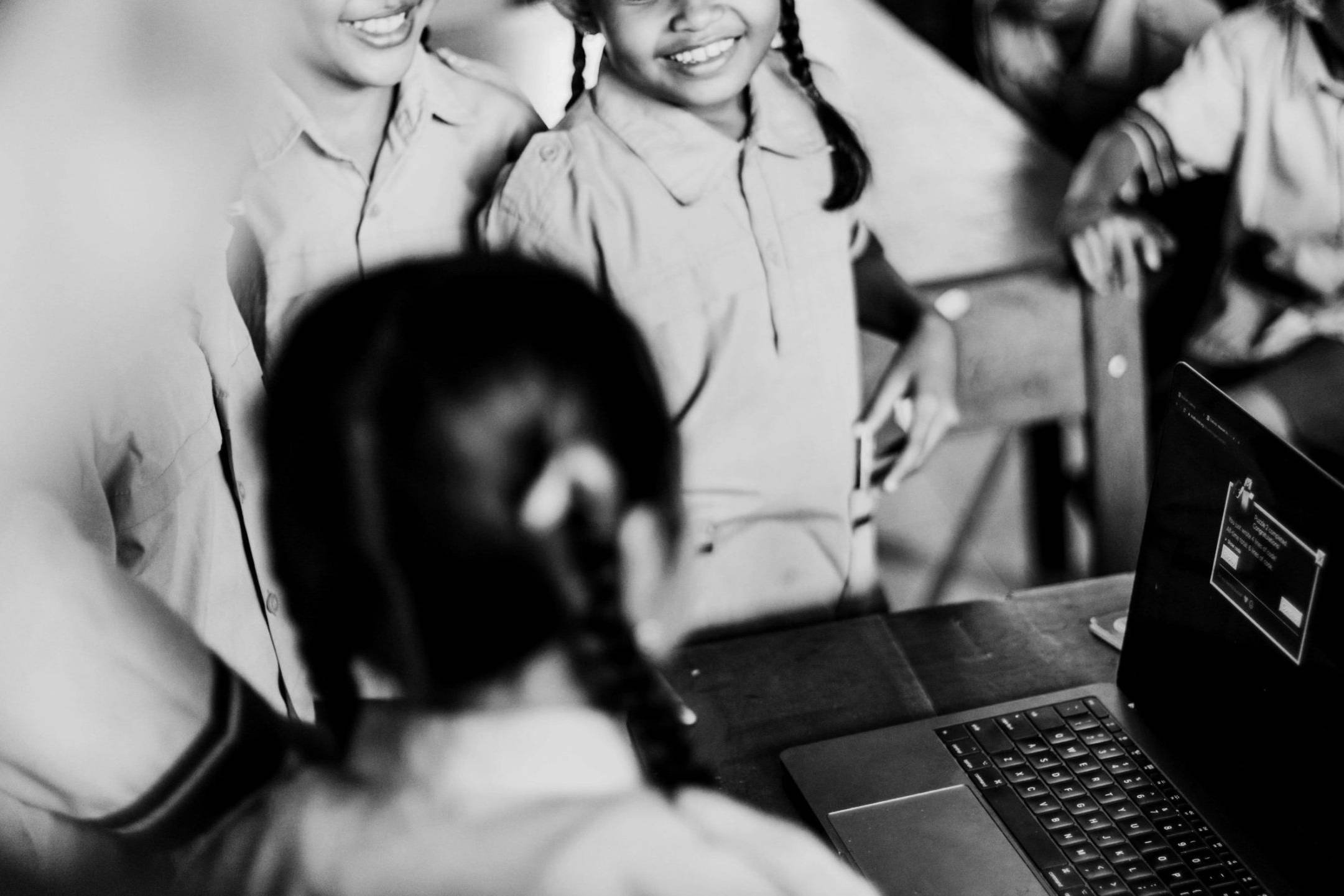 Three smiling children in school uniforms gather around a laptop in a classroom. The mood is joyful and engaging, suggesting learning and collaboration.