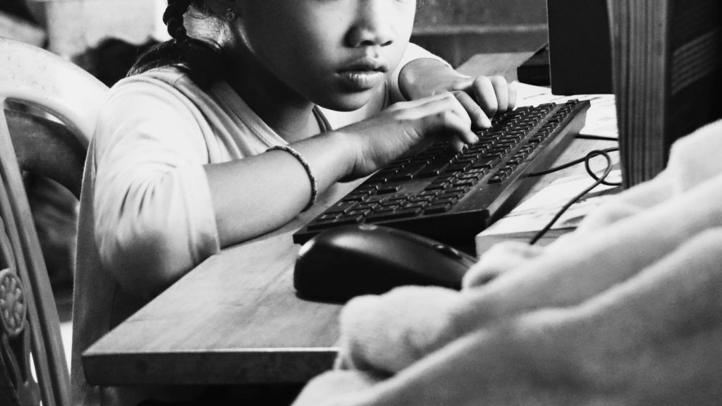 A young girl intently uses a desktop computer, her fingers poised on the keyboard. Black-and-white tones evoke a focused, contemplative mood.