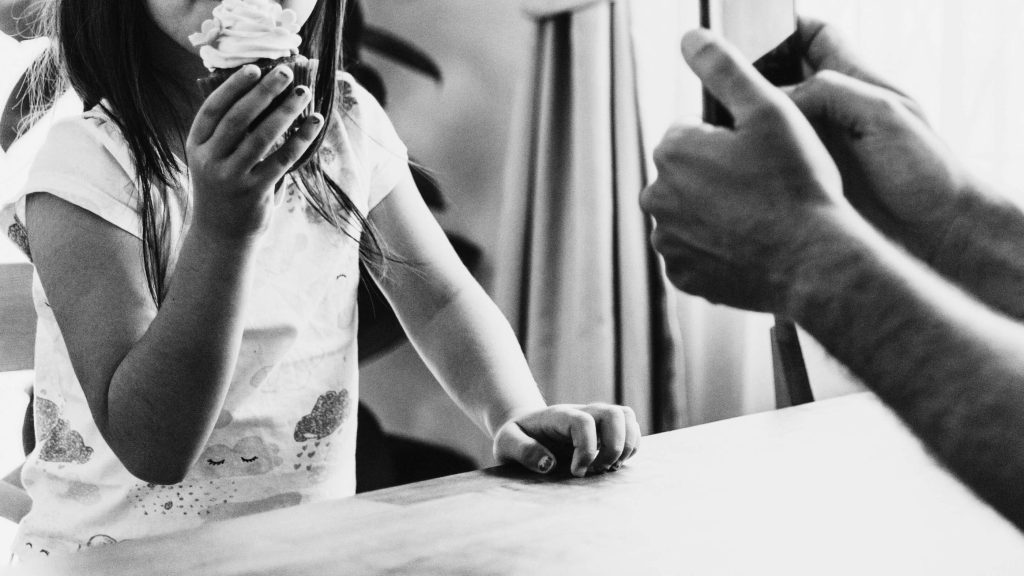 A black-and-white image features a child in a printed shirt holding a cupcake topped with swirled icing. An adult holds a phone, likely taking a photo.