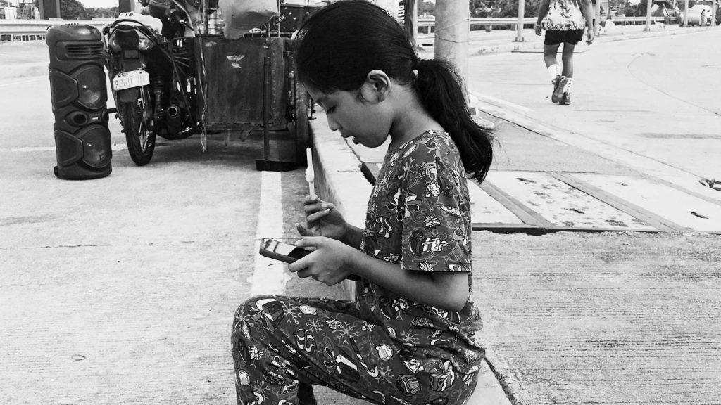 A young girl in patterned pajamas sits on a curb, using a smartphone and holding a toothbrush. A tricycle and loudspeaker are in the background. The scene is in black and white, conveying a contemplative mood.