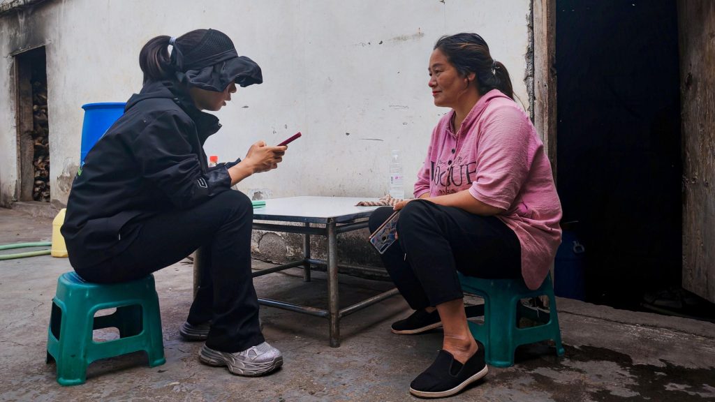 Two women sit on small stools outside, engaged in conversation. One wears black, holding a phone; the other is in pink, listening attentively. The tone is candid and relaxed.