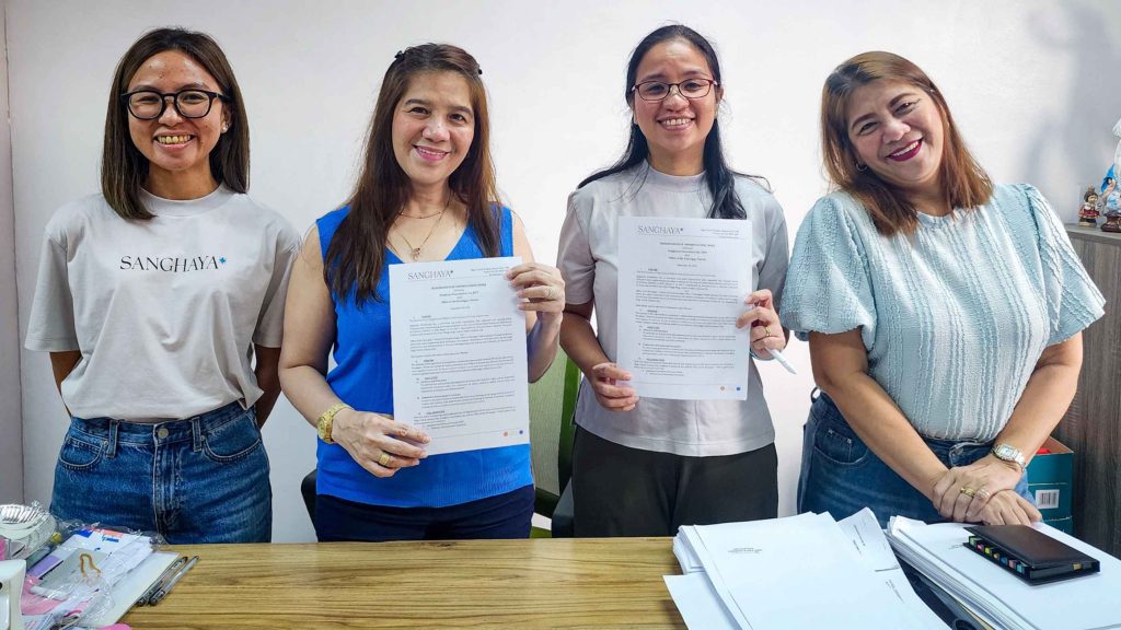 Four smiling women stand behind a table. Two hold documents. The setting is a bright office, conveying a positive and professional atmosphere.