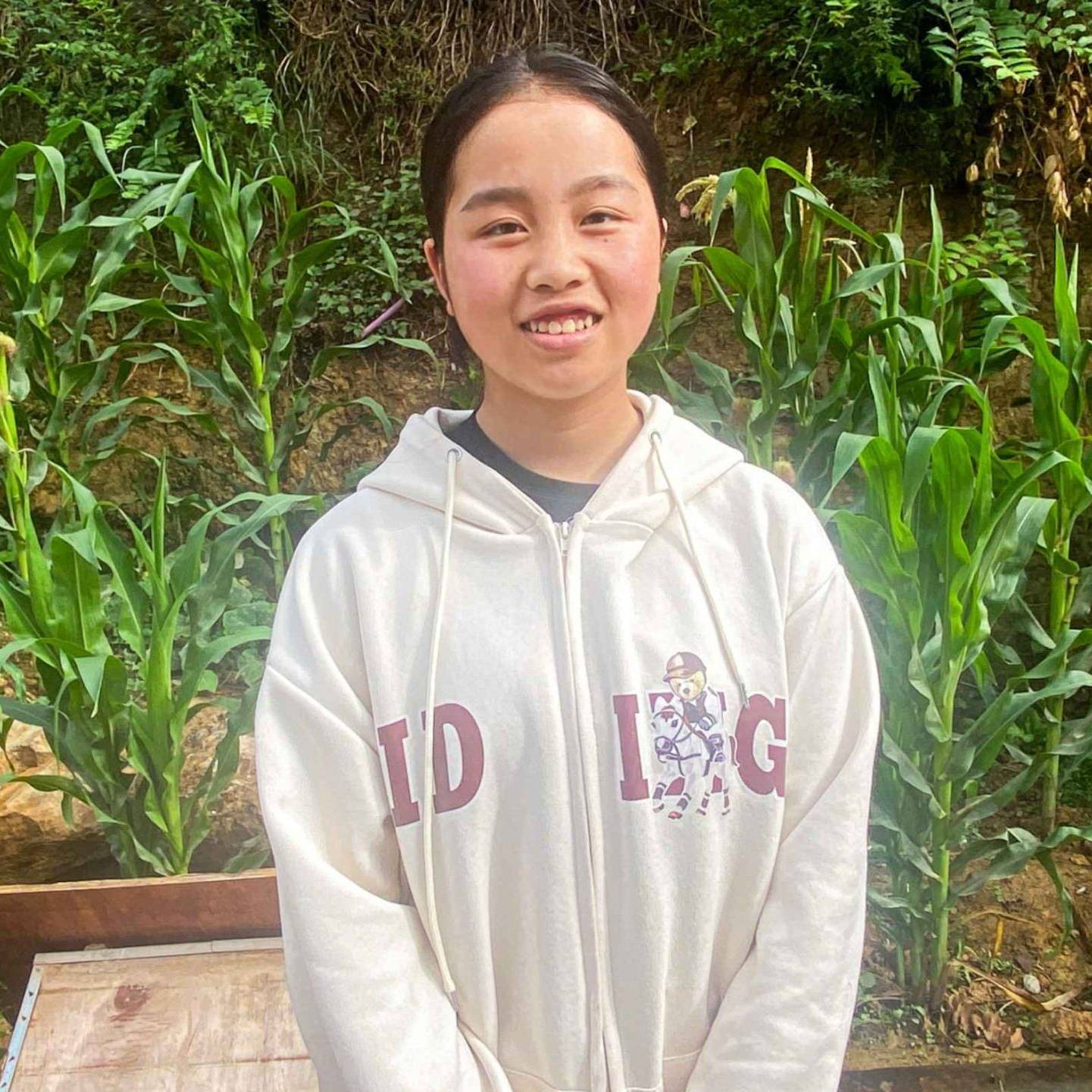 A young person stands smiling in a lush garden with tall green plants, likely corn. They wear a light hoodie with letters and a cartoon character. The atmosphere is serene and natural.