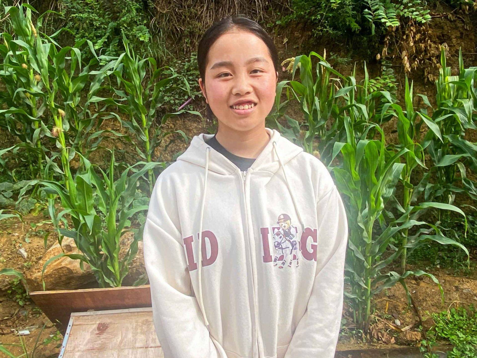 A young person stands smiling in a lush garden with tall green plants, likely corn. They wear a light hoodie with letters and a cartoon character. The atmosphere is serene and natural.
