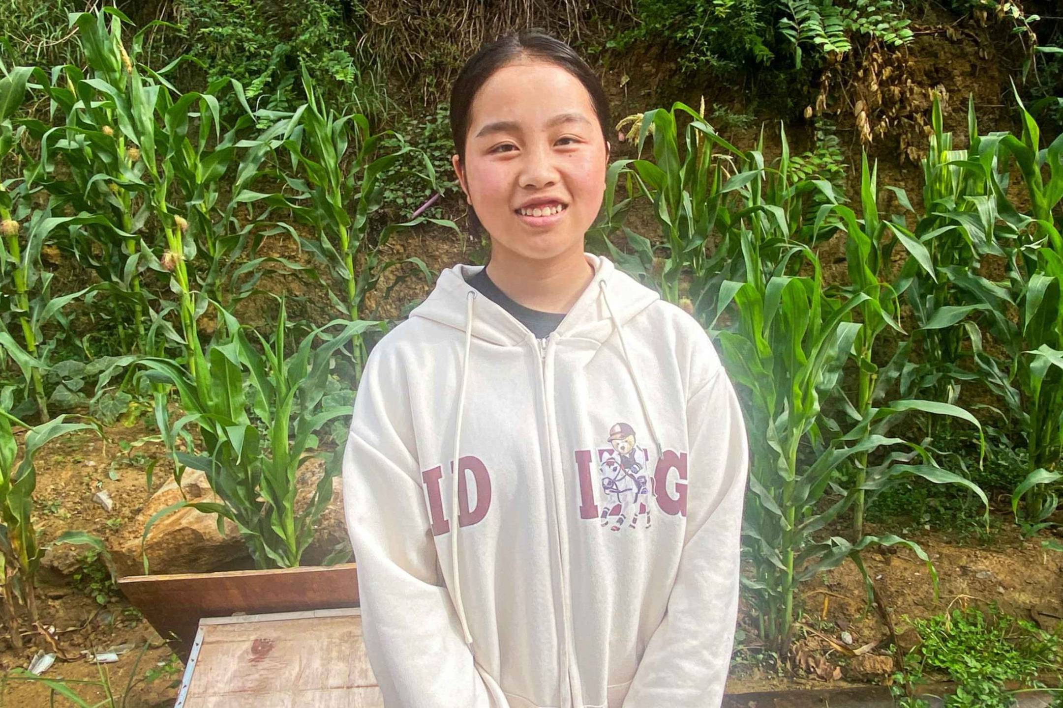 A young person stands smiling in a lush garden with tall green plants, likely corn. They wear a light hoodie with letters and a cartoon character. The atmosphere is serene and natural.