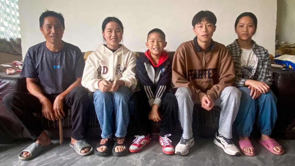 A family of five sits on a couch in a relaxed, casual setting. They are smiling and wearing casual clothing, exuding warmth and togetherness.
