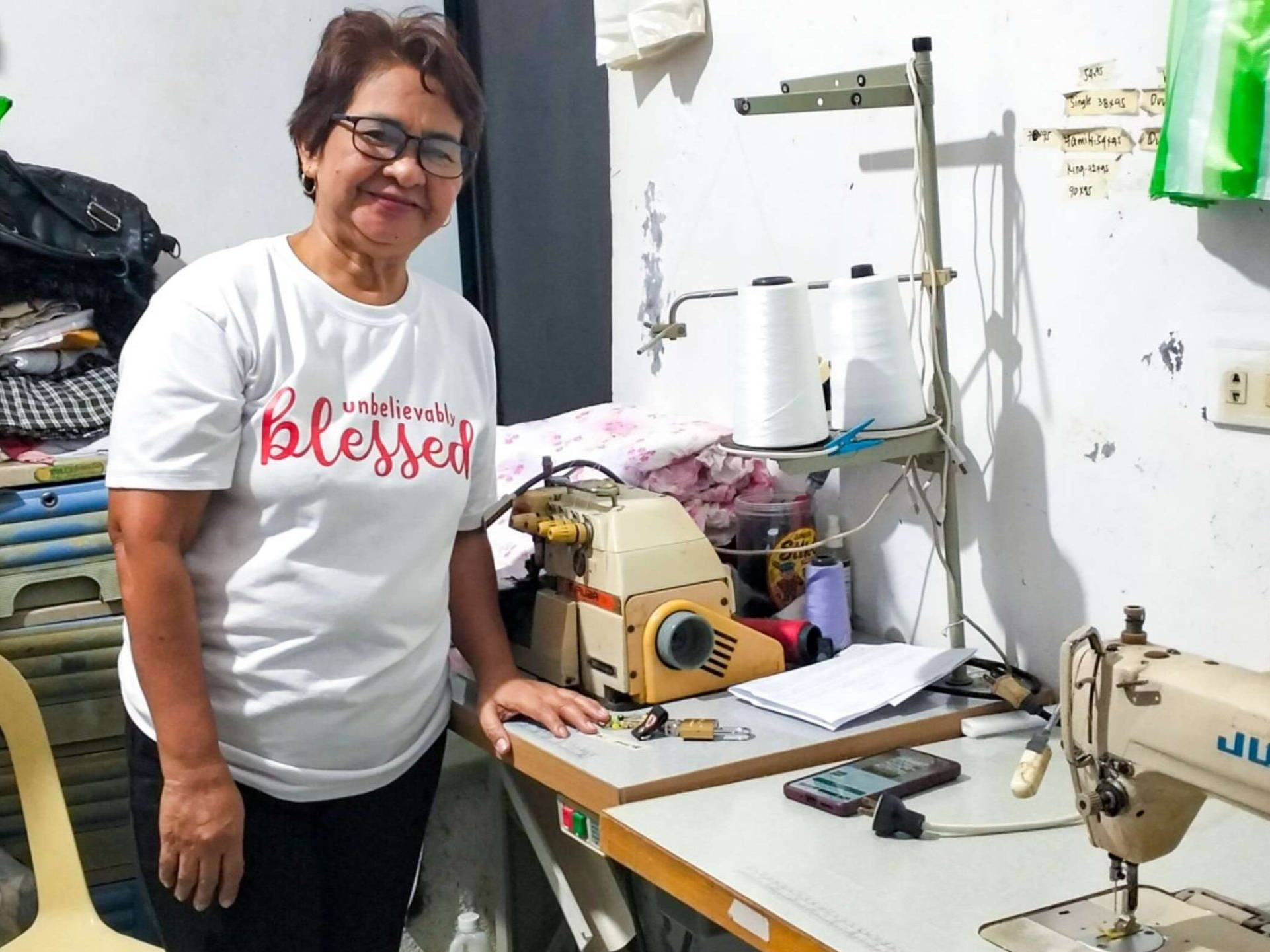 Woman smiling beside sewing machines in a cluttered room, wearing a white T-shirt. The atmosphere is warm and industrious.