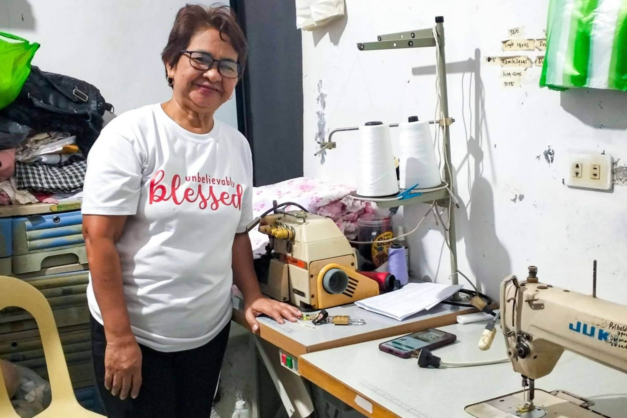 Woman smiling beside sewing machines in a cluttered room, wearing a white T-shirt. The atmosphere is warm and industrious.