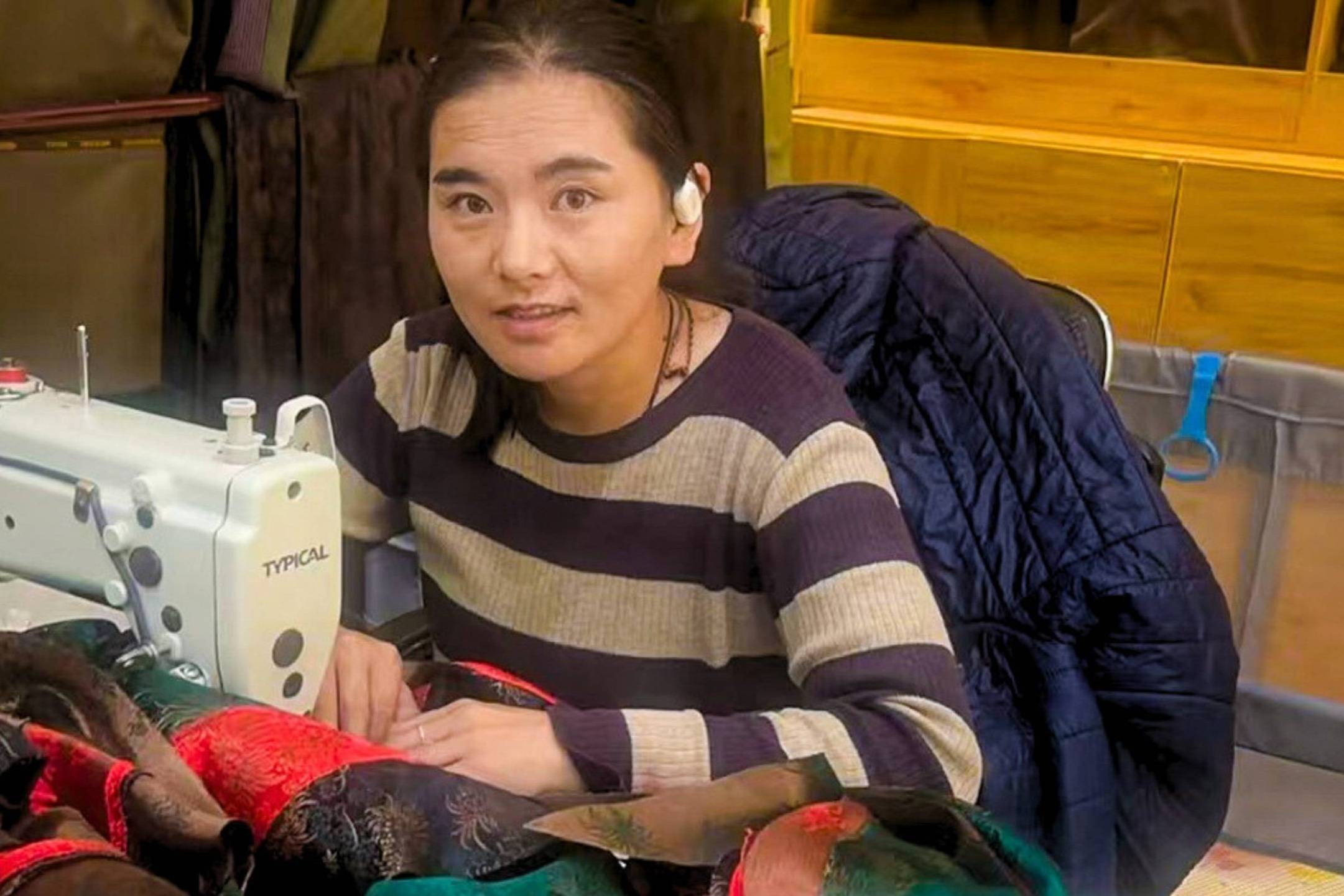 A woman sits at a sewing machine, surrounded by colorful fabric. She wears a striped sweater, conveying focus and determination.