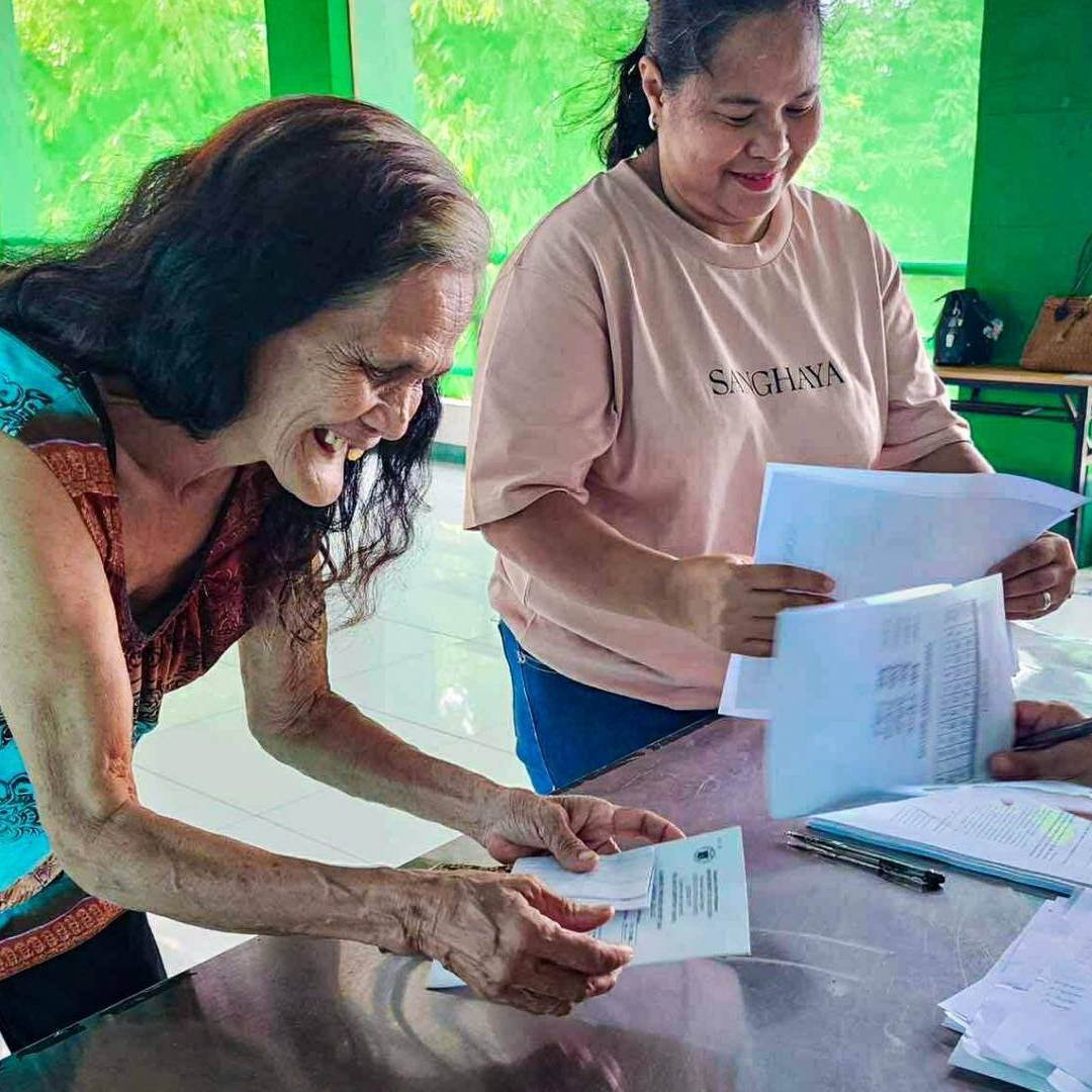 An elderly woman with long hair smiles broadly as she receives documents from a seated person at a table. A second woman stands nearby, holding papers. The scene appears joyful and welcoming, with several other people in the background engaged in paperwork.