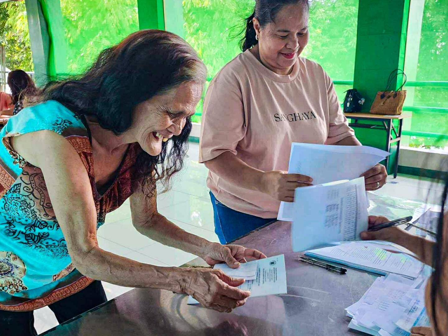 An elderly woman with long hair smiles broadly as she receives documents from a seated person at a table. A second woman stands nearby, holding papers. The scene appears joyful and welcoming, with several other people in the background engaged in paperwork.