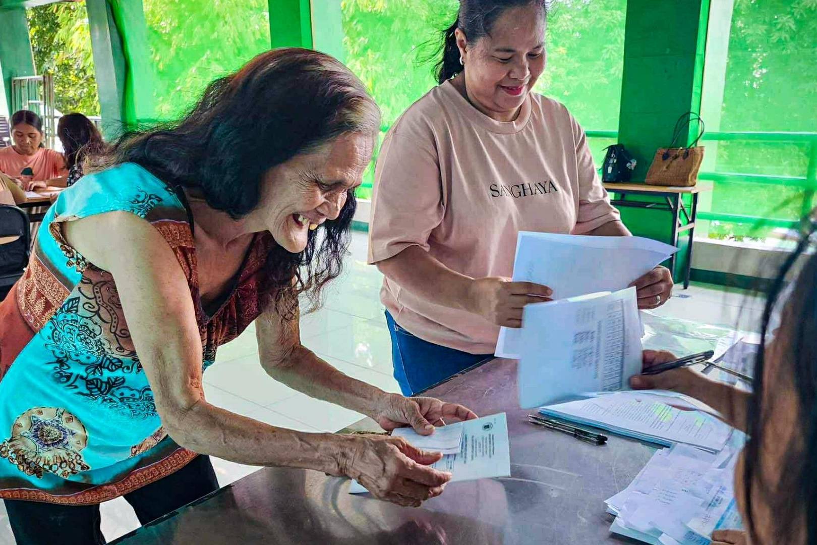 An elderly woman with long hair smiles broadly as she receives documents from a seated person at a table. A second woman stands nearby, holding papers. The scene appears joyful and welcoming, with several other people in the background engaged in paperwork.