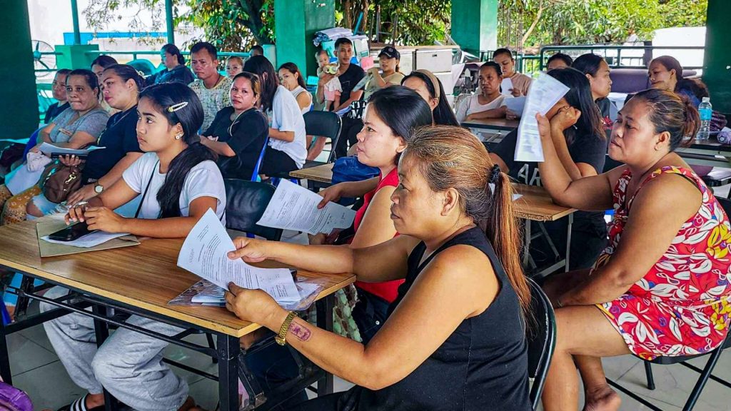 A group of adults attentively listens in a classroom setting, holding documents. The diverse group appears engaged, suggesting a serious and focused atmosphere.
