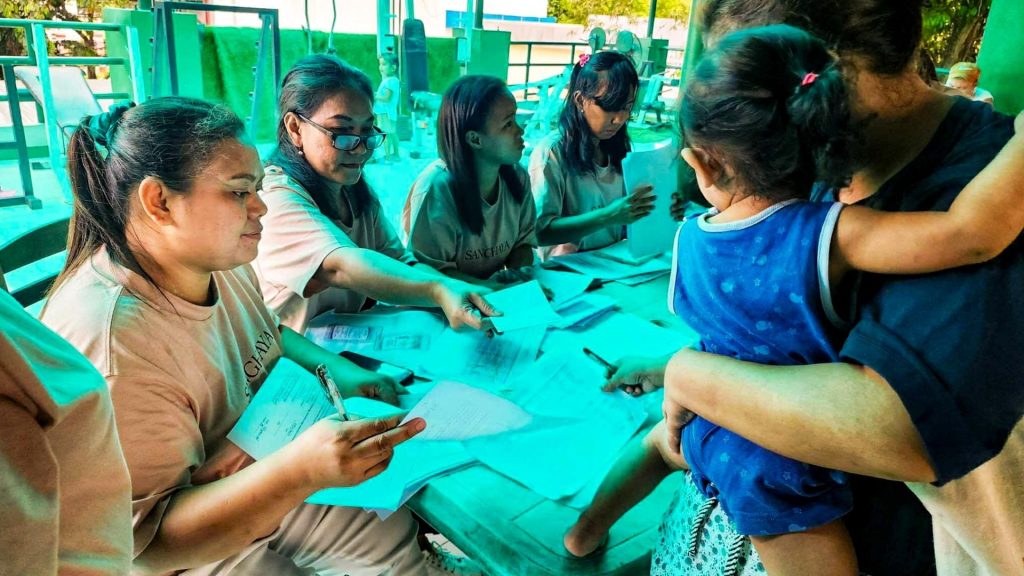 A group of people wearing light shirts sit at a table, actively engaged in paperwork, while a person holding a child approaches them. The setting appears busy and collaborative.