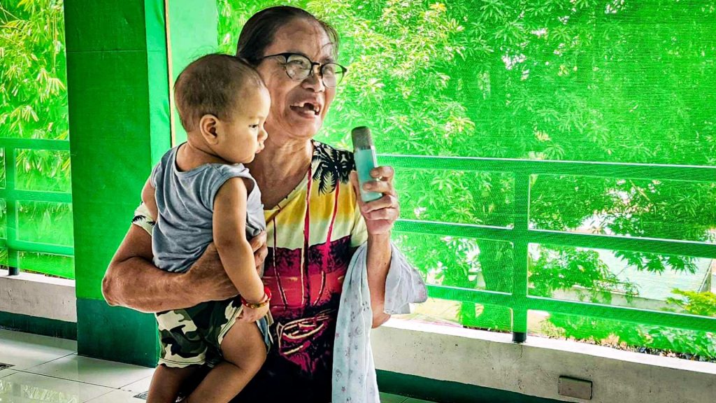 Elderly woman in glasses joyfully sings into a microphone while holding a child. They stand on a balcony with lush greenery in the background.