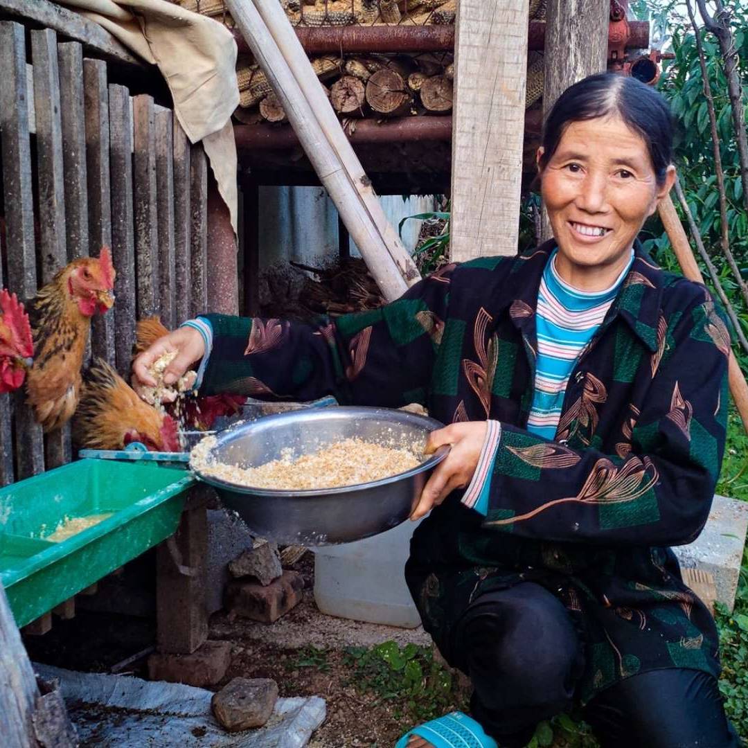 A smiling person kneels outdoors, feeding chickens from a bowl. They're next to a wooden coop, with green plants in the background, conveying a rural setting.