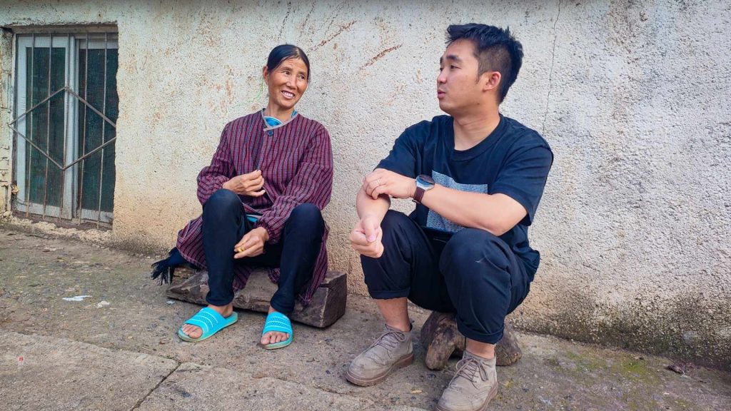 A man and woman are sitting on a sidewalk in front of a textured wall. The woman, wearing a striped jacket and blue sandals, smiles warmly. The man, in a black shirt and pants, sits with a relaxed posture, engaged in conversation. The scene exudes a sense of casual, friendly interaction.
