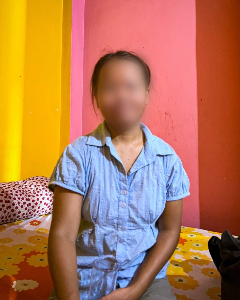 A person in a light blue shirt sits on a colorful bed, with vibrant yellow and pink walls in the background. Her face blurred for privacy. The setting is warm and inviting.