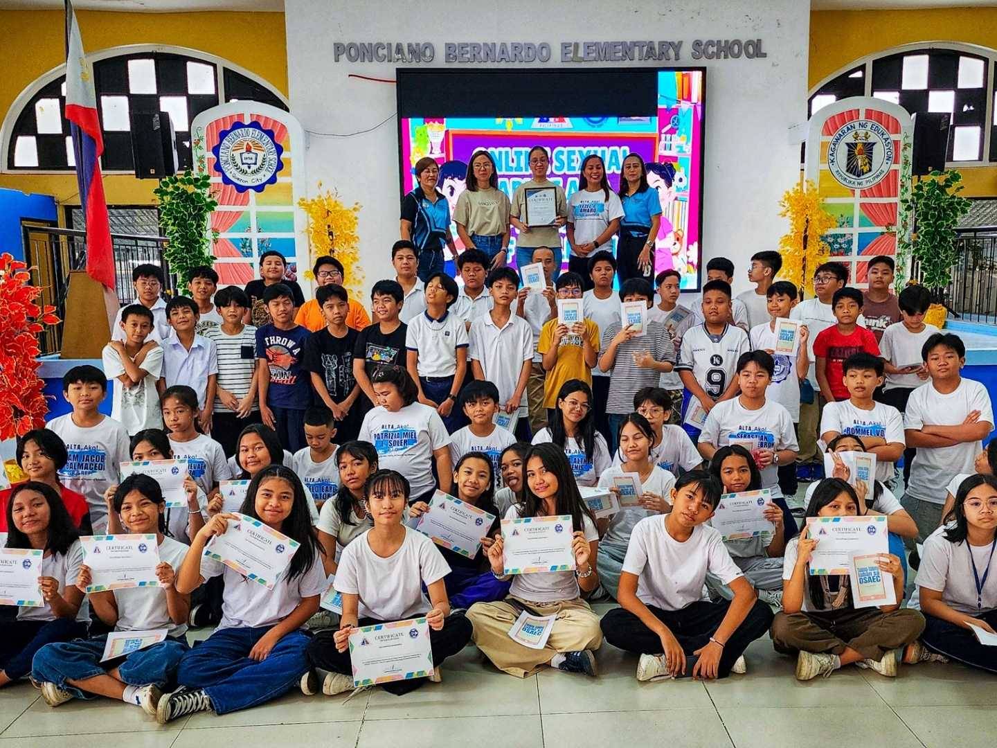 A group of students at Ponciiano Bernardo Elementary School proudly display certificates in a colorful auditorium, smiling and celebrating their achievements.