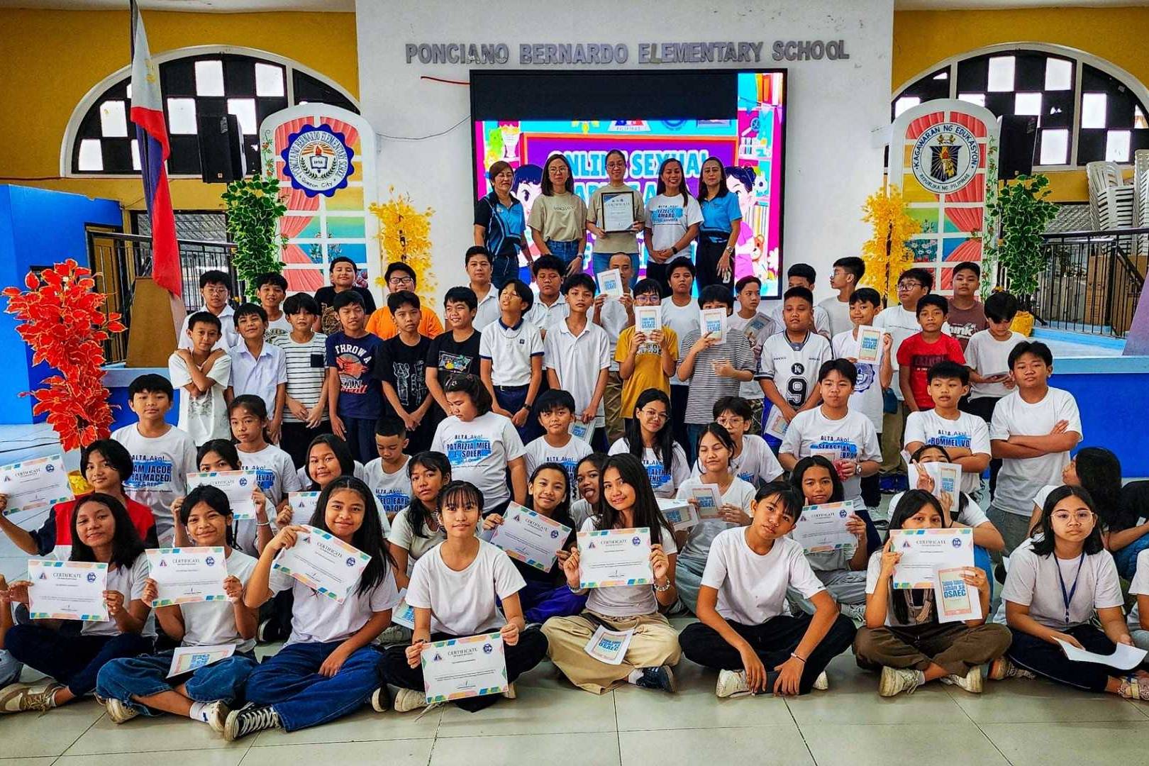 A group of students at Ponciiano Bernardo Elementary School proudly display certificates in a colorful auditorium, smiling and celebrating their achievements.