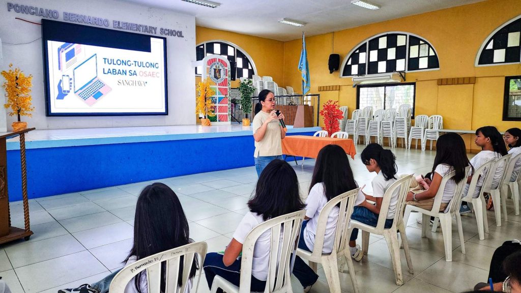 A woman speaks to seated students in a school hall. A blue stage with a screen displays "Tulong-Tulong Laban Sa OSAEC." The ambiance is educational and attentive.